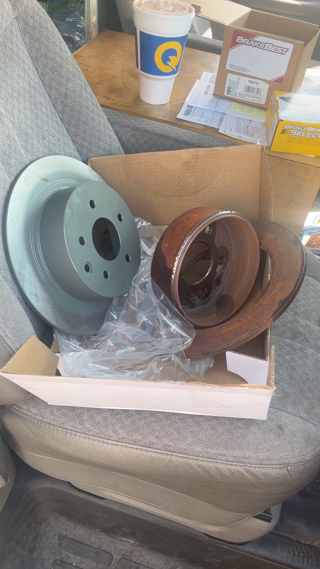 A new brake rotor beside rusty old rotors in a box on a vehicle seat
